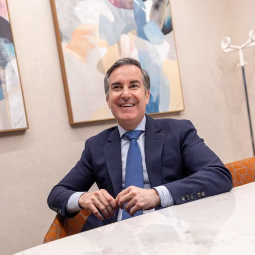 Hombre con traje azul marino y corbata azul claro sentado ante una mesa blanca, con las manos entrelazadas; detrás hay dos cuadros abstractos y un perchero blanco en una sala de estilo moderno.