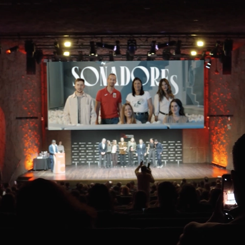 Vista desde el público de un escenario de teatro durante un evento. Una gran pantalla central muestra la palabra 'SOÑADORES' junto a las fotos de seis personas. En el escenario, un grupo de ponentes permanece de pie mientras un presentador habla desde un atril lateral, bajo una iluminación cálida y profesional.