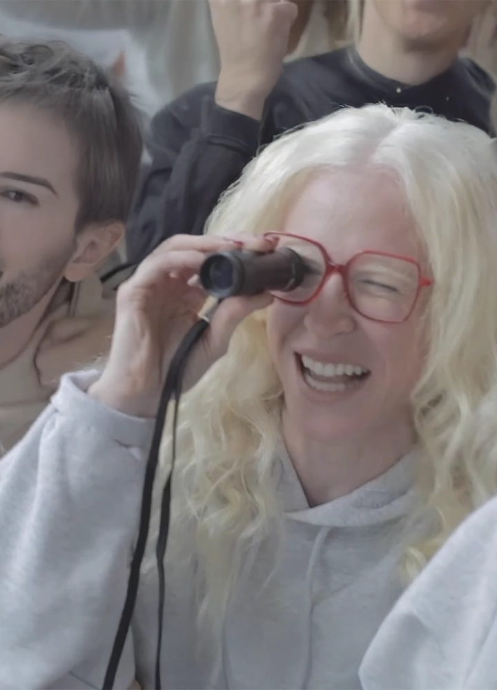 Primer plano de una mujer con el cabello rubio platino y gafas de montura roja, que sonríe con entusiasmo mientras utiliza un monocular de mano para ver a lo lejos. Viste una sudadera gris clara y se encuentra rodeada de otras personas en un ambiente concurrido.