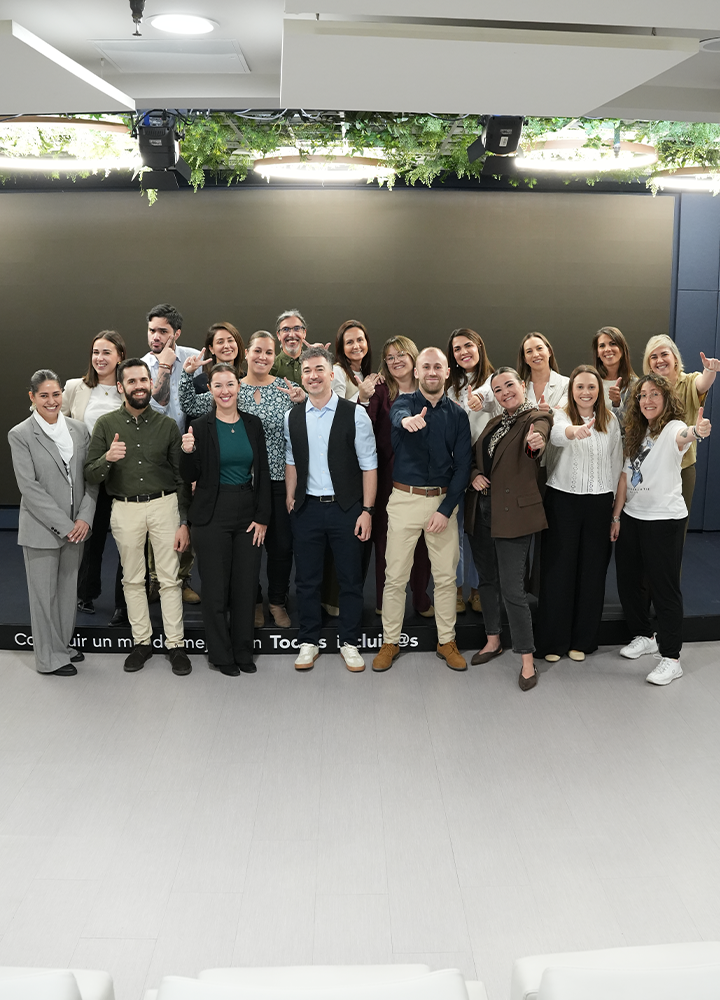 Grupo numeroso de personas adultas posando de pie en varias filas dentro de un espacio interior corporativo. Todas están alineadas frente a un fondo oscuro liso, con parte inferior en color negro y techo con iluminación integrada y vegetación colgante. La vestimenta es profesional y semiformal, con trajes, americanas, camisas, blusas y pantalones de vestir en tonos neutros. Varias personas levantan el pulgar en gesto positivo. Los rostros aparecen desenfocados. El suelo es claro y liso, y el conjunto transmite un ambiente de equipo y encuentro profesional.