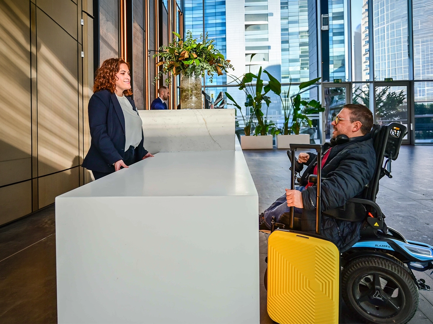 Mostrador de recepción blanco en un vestíbulo moderno y amplio, con suelo oscuro y grandes ventanales que dejan ver edificios altos en el exterior. Frente al mostrador, una persona usuaria de silla de ruedas eléctrica se sitúa a la altura del mostrador adaptado y sostiene una maleta rígida amarilla con asa extendida. Detrás del mostrador, otra persona permanece de pie atendiendo. Al fondo se observan plantas decorativas, otra persona junto a un segundo mostrador y una entrada acristalada. La iluminación es natural y el espacio transmite un entorno accesible y corporativo.