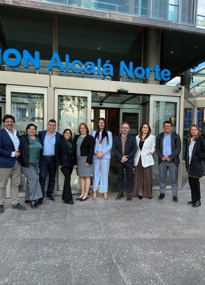 Grupo de personas adultas posando en fila en el exterior de un edificio moderno. Todas están de pie, alineadas frente a una entrada acristalada con puertas correderas. Sobre la entrada, un rótulo azul con el texto “ILUNION Alcalá Norte”. La vestimenta es profesional y semiformal, con trajes, americanas, abrigos y pantalones de vestir. El suelo es de piedra gris y la fachada combina cristal y elementos metálicos. Los rostros aparecen desenfocados y la escena transmite un contexto corporativo.
