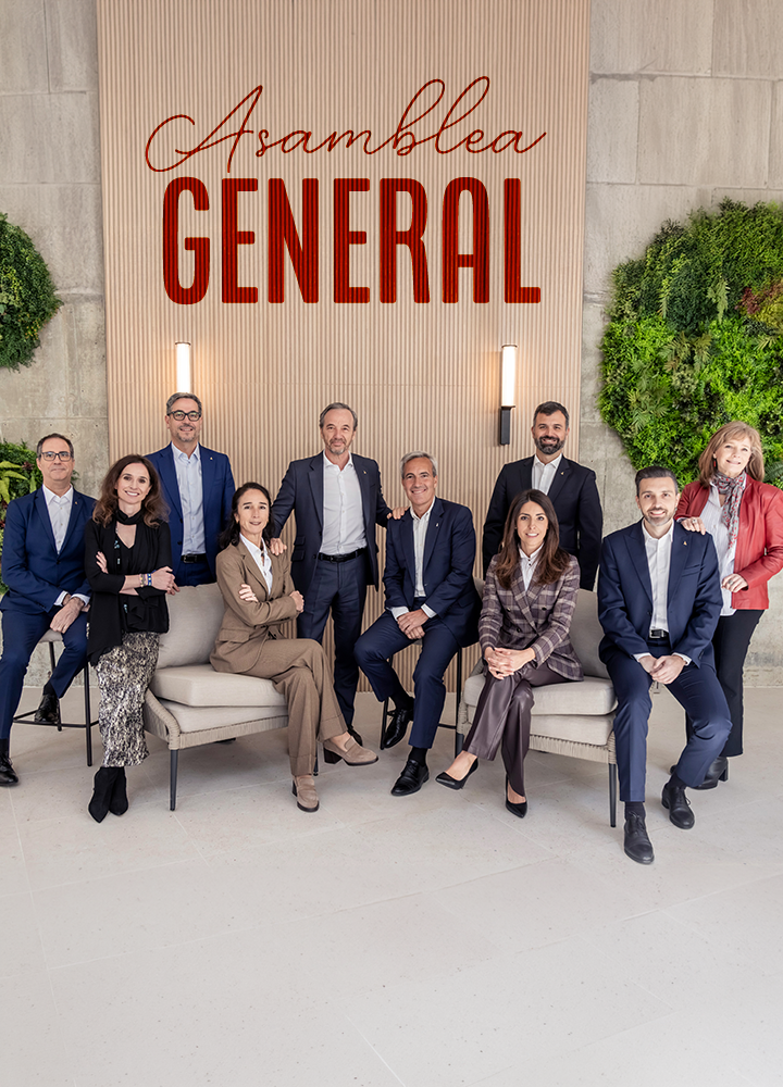 Grupo de personas adultas posando juntas en un entorno interior de estilo corporativo, algunas sentadas en sillones claros y otras de pie detrás. Todas visten ropa formal y semiformal, como trajes, americanas y conjuntos de vestir; los rostros aparecen desenfocados. Al fondo, una pared decorativa combina paneles de piedra clara, listones verticales y zonas de vegetación verde a ambos lados. En la parte superior central destaca un gran rótulo con el texto “Asamblea GENERAL”, con “Asamblea” en tipografía manuscrita y “GENERAL” en letras grandes y rojas. La iluminación es uniforme y el conjunto transmite un ambiente institucional y profesional.