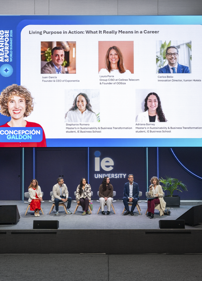 Vista general del mismo evento en un auditorio de IE University. En primer plano se ve el escenario con siete personas sentadas en fila participando en una mesa redonda. En el fondo, una gran pantalla muestra los retratos y nombres de las personas participantes, junto con el título “Living Purpose in Action: What It Really Means in a Career” y el logotipo de IE University. El escenario es amplio, con elementos decorativos minimalistas y luz azul alrededor de la pantalla.