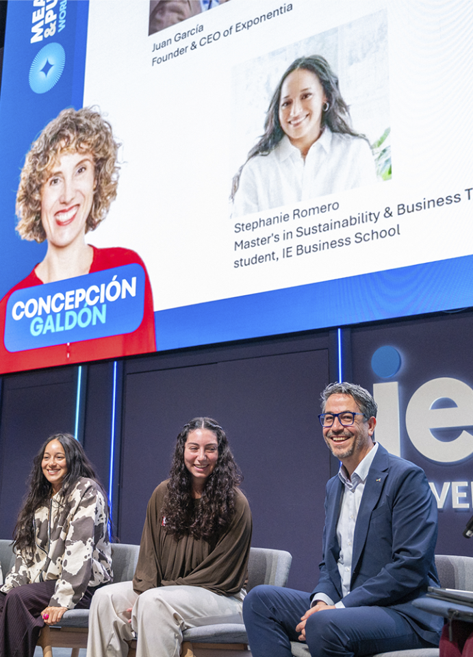 Escenario de un auditorio de IE University durante un evento. En primer plano, tres personas están sentadas en sillas como parte de un panel. En el fondo, una gran pantalla muestra una diapositiva con retratos y texto informativo. Se leen nombres y cargos, entre ellos “Juan García, Founder & CEO of Exponentia” y “Stephanie Romero, Master’s in Sustainability & Business Transformation student, IE Business School”. A la izquierda de la pantalla aparece una ilustración con el nombre “Concepción Galdón”. El escenario tiene iluminación profesional y diseño moderno en tonos azules y oscuros.