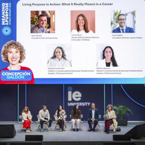 Vista general del mismo evento en un auditorio de IE University. En primer plano se ve el escenario con siete personas sentadas en fila participando en una mesa redonda. En el fondo, una gran pantalla muestra los retratos y nombres de las personas participantes, junto con el título “Living Purpose in Action: What It Really Means in a Career” y el logotipo de IE University. El escenario es amplio, con elementos decorativos minimalistas y luz azul alrededor de la pantalla.
