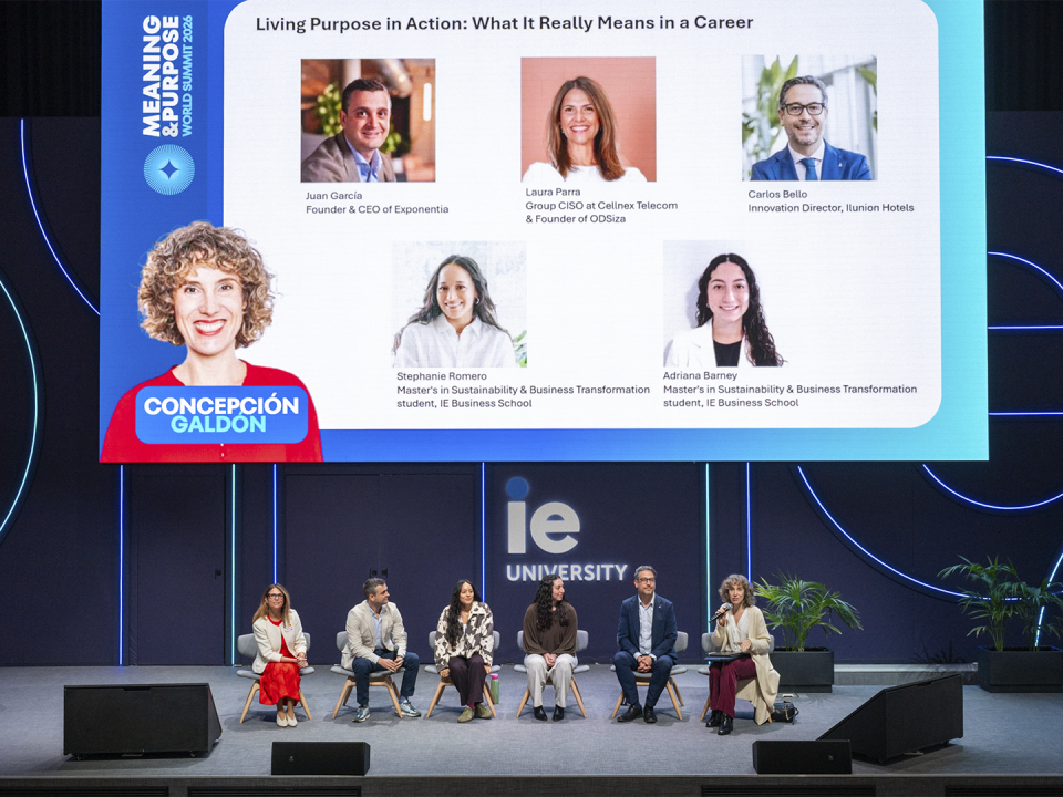 Vista general del mismo evento en un auditorio de IE University. En primer plano se ve el escenario con siete personas sentadas en fila participando en una mesa redonda. En el fondo, una gran pantalla muestra los retratos y nombres de las personas participantes, junto con el título “Living Purpose in Action: What It Really Means in a Career” y el logotipo de IE University. El escenario es amplio, con elementos decorativos minimalistas y luz azul alrededor de la pantalla.