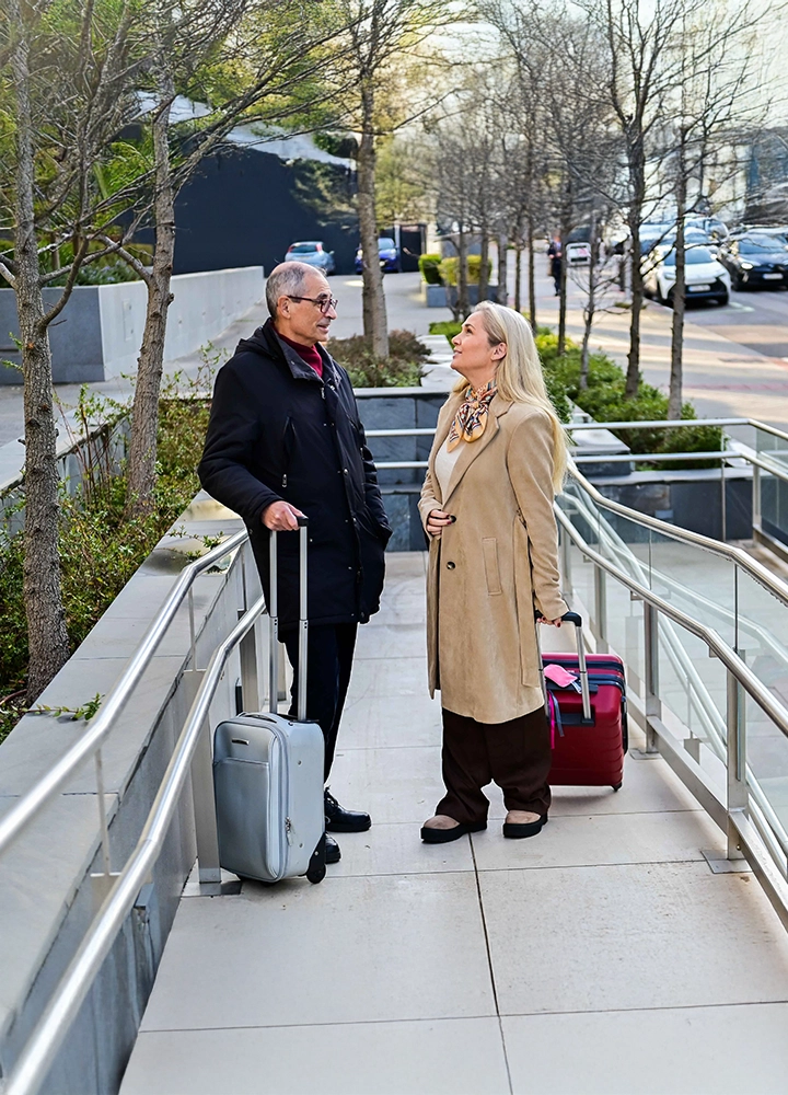 Dos personas se encuentran de pie en una rampa urbana con barandillas a ambos lados. Cada persona lleva una maleta con ruedas. La rampa está rodeada de árboles y vegetación, junto a una acera y una calle con coches al fondo. El entorno es exterior y parece un acceso adaptado a un edificio.