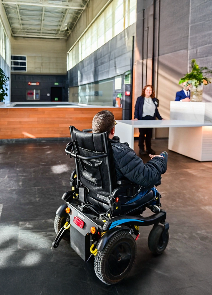 Una persona se desplaza en una silla de ruedas eléctrica dentro del vestíbulo moderno de un edificio. Al frente hay un mostrador de recepción blanco con una planta encima y dos personas de pie detrás del mostrador. El espacio es amplio, con suelo pulido, paredes altas y grandes ventanales que dejan entrar luz natural.