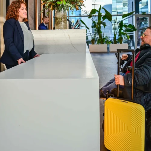 Una persona en silla de ruedas eléctrica se encuentra frente a un mostrador de recepción blanco en el interior de un edificio moderno. La persona sostiene el asa de una maleta amarilla colocada a su lado. Al otro lado del mostrador hay otra persona de pie. El espacio es amplio, con plantas decorativas, paredes altas y grandes ventanales que dejan pasar la luz natural.
