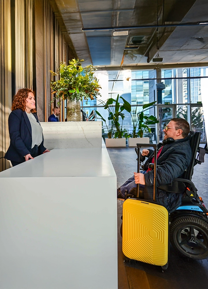 Una persona en silla de ruedas eléctrica se encuentra frente a un mostrador de recepción blanco en el interior de un edificio moderno. La persona sostiene el asa de una maleta amarilla colocada a su lado. Al otro lado del mostrador hay otra persona de pie. El espacio es amplio, con plantas decorativas, paredes altas y grandes ventanales que dejan pasar la luz natural.
