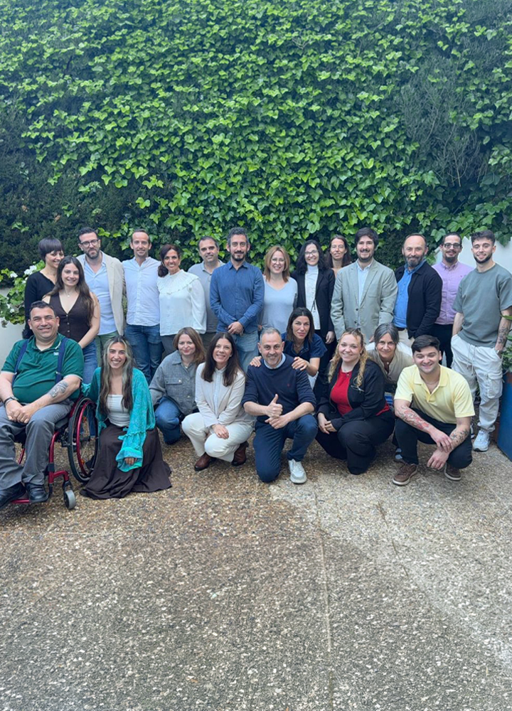 Grupo numeroso de personas adultas posando juntas al aire libre, organizadas en varias filas, algunas de pie y otras agachadas. El fondo está cubierto por una pared vegetal densa de color verde. La vestimenta es variada, combinando ropa profesional e informal. En primer plano se ve un suelo de grava clara. Los rostros aparecen desenfocados.