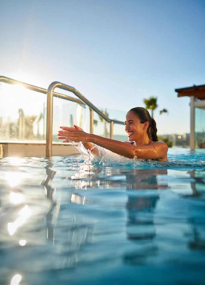 Piscina exterior a nivel del agua, con una persona apoyada en el borde y los brazos extendidos, salpicando el agua. Barandilla metálica a la izquierda, reflejos del sol sobre la superficie y cielo despejado al fondo, con edificios y palmeras lejanas.