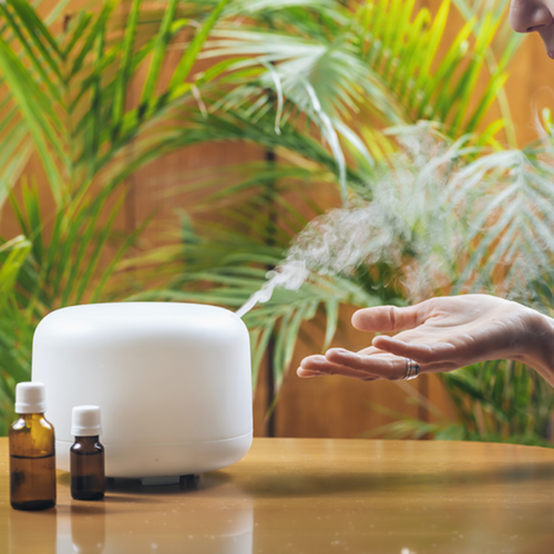 Una persona acerca su mano a un difusor de aceites esenciales blanco que emite vapor suave sobre una mesa de madera. Junto al difusor hay dos pequeños frascos de vidrio oscuro. Al fondo se ven plantas verdes, creando un ambiente tranquilo y natural.