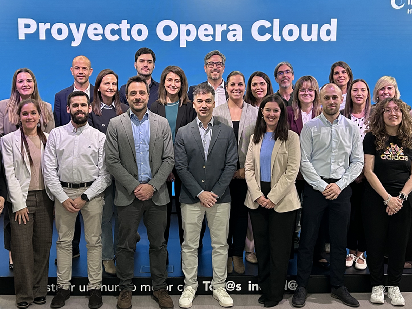 Grupo numeroso de personas adultas posando de pie en varias filas en un entorno corporativo interior. Al fondo, una gran pantalla azul ocupa todo el ancho de la imagen con el texto blanco “Proyecto Opera Cloud” en la parte superior. Las personas visten ropa profesional y semiformal, como americanas, camisas, blusas y pantalones de vestir; algunas combinan prendas más informales. Todas están orientadas hacia la cámara, con postura recta y manos juntas delante del cuerpo o a los lados. Los rostros aparecen desenfocados y la iluminación es uniforme y frontal.