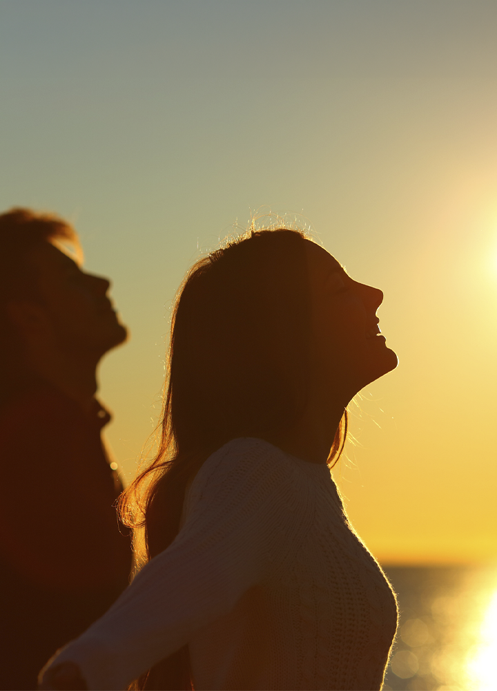 Dos personas adultas vistas de perfil y a contraluz, situadas en primer plano frente al mar. Las figuras aparecen en silueta, con los rostros desenfocados, iluminadas por la luz cálida de un atardecer. El sol bajo se refleja sobre el agua, creando un fondo dorado y un cielo despejado con tonos amarillos y anaranjados. La escena transmite calma y amplitud del entorno costero.