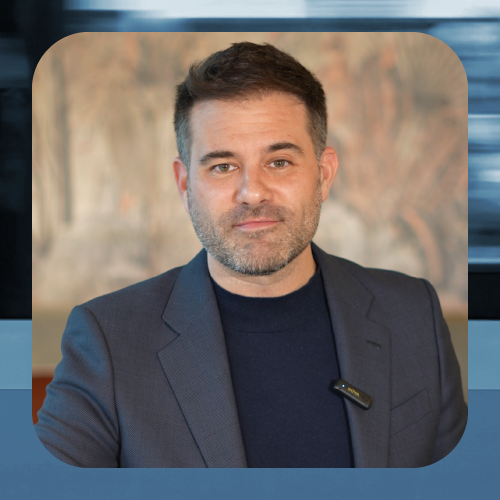 Retrato de una persona adulta en plano medio, centrada en la imagen, con el rostro difuminado. Viste una chaqueta oscura sobre una camiseta oscura y lleva un pequeño micrófono de solapa sujeto a la ropa. El fondo es interior y desenfocado, con una pintura o mural en tonos cálidos que aporta textura visual. La imagen está enmarcada con bordes redondeados y un marco gráfico en tonos azules, lo que sugiere un contexto profesional, institucional o de comunicación corporativa.