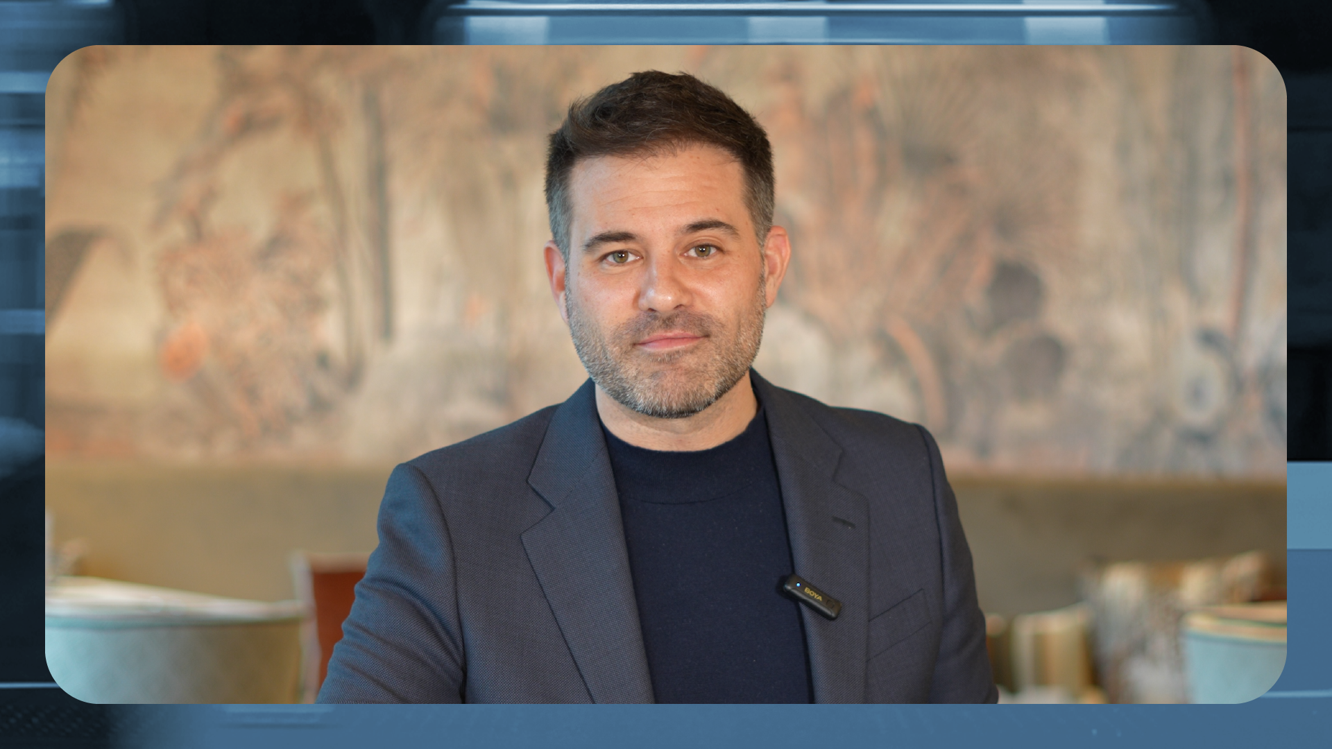 Retrato de una persona adulta en plano medio, centrada en la imagen, con el rostro difuminado. Viste una chaqueta oscura sobre una camiseta oscura y lleva un pequeño micrófono de solapa sujeto a la ropa. El fondo es interior y desenfocado, con una pintura o mural en tonos cálidos que aporta textura visual. La imagen está enmarcada con bordes redondeados y un marco gráfico en tonos azules, lo que sugiere un contexto profesional, institucional o de comunicación corporativa.