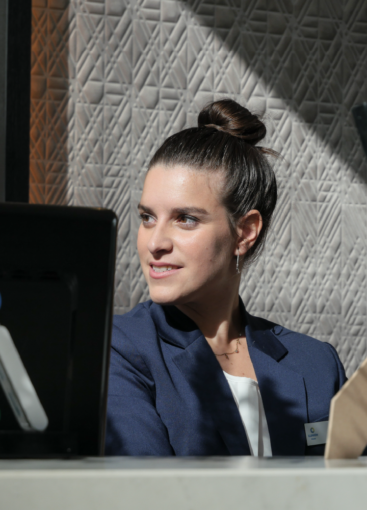 Persona sentada detrás de un mostrador o mesa de trabajo, vestida con una chaqueta azul sobre una prenda clara. El cabello está recogido en un moño alto. Frente a la persona hay un monitor negro y, a un lado, un objeto de color claro con forma rectangular. El fondo es una pared con relieve geométrico en tonos grises, iluminada por luz lateral que crea sombras marcadas y un ambiente profesional e interior.