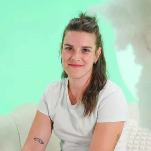 Persona sentada en un sillón claro, con el cuerpo ligeramente girado hacia un lado. Viste una camiseta blanca de manga corta y lleva el cabello recogido en la parte superior. El fondo es un escenario de estudio con iluminación suave y un degradado en tonos verdes, acompañado de formas difusas que aportan un ambiente tranquilo y cuidado.