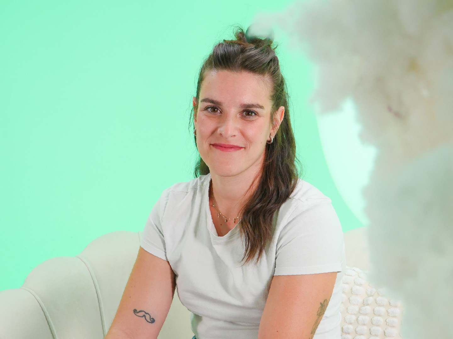 Persona sentada en un sillón claro, con el cuerpo ligeramente girado hacia un lado. Viste una camiseta blanca de manga corta y lleva el cabello recogido en la parte superior. El fondo es un escenario de estudio con iluminación suave y un degradado en tonos verdes, acompañado de formas difusas que aportan un ambiente tranquilo y cuidado.