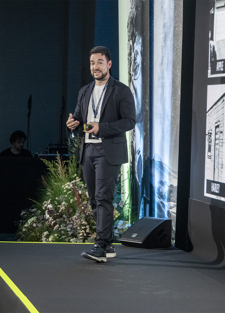 Persona presentadora caminando por un escenario durante una charla, sosteniendo un pequeño mando en la mano. Lleva ropa formal y una acreditación. A su lado hay una pantalla vertical con contenido gráfico y, al fondo, decoración con plantas.