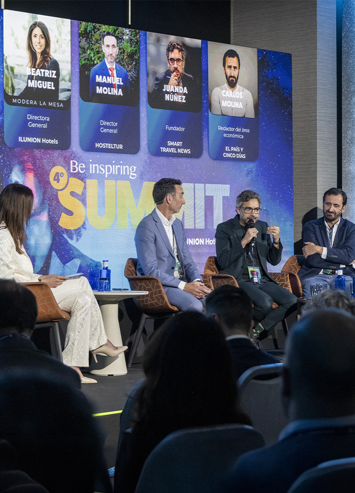 Conversación en un escenario con varias personas sentadas, una de ellas hablando con un micrófono en la mano. La audiencia aparece en primer plano. Al fondo se ve una pantalla grande con el título “Be inspiring Summit” y fotos de los participantes.