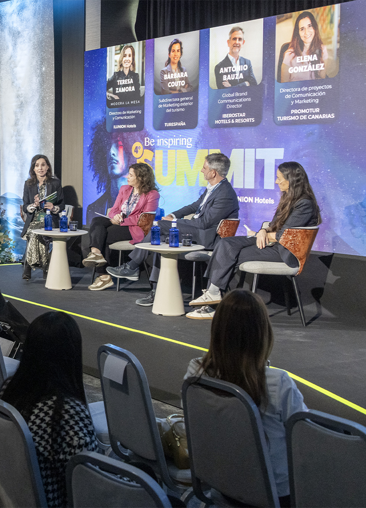 Panel de discusión en un escenario con varias personas sentadas en sillas modernas. Una persona habla mientras otras escuchan. Detrás hay una gran pantalla con fotos, nombres y cargos de participantes del evento “Summit”.