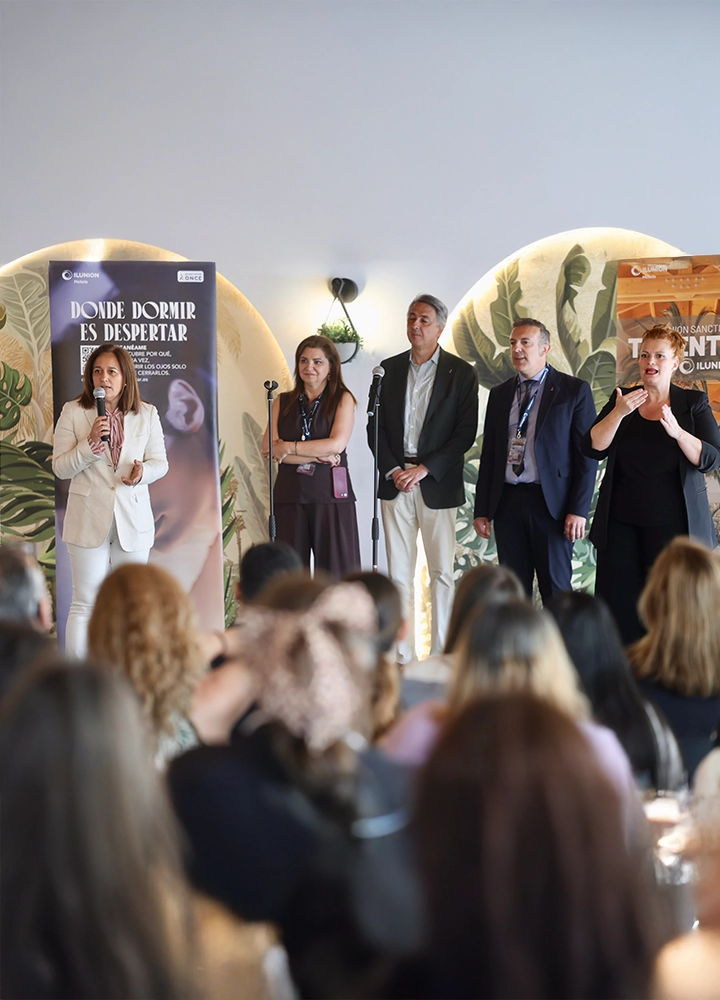 En un espacio interior, varias personas están de pie sobre un pequeño escenario frente a un público sentado. Delante del escenario hay micrófonos y detrás un fondo decorado con motivos vegetales y paneles con texto. El público aparece desenfocado en primer plano.