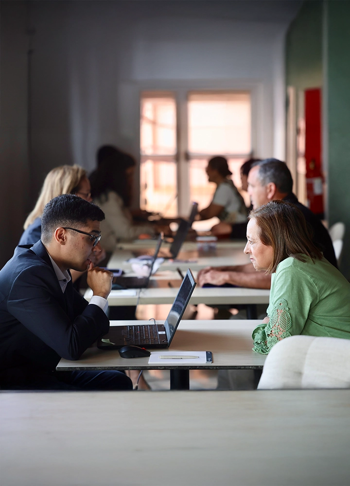 Varias personas se encuentran sentadas en mesas largas dentro de una sala luminosa. En primer plano, dos personas se sientan frente a frente con un ordenador portátil abierto entre ellas, mientras otras parejas trabajan en mesas similares al fondo de la sala.