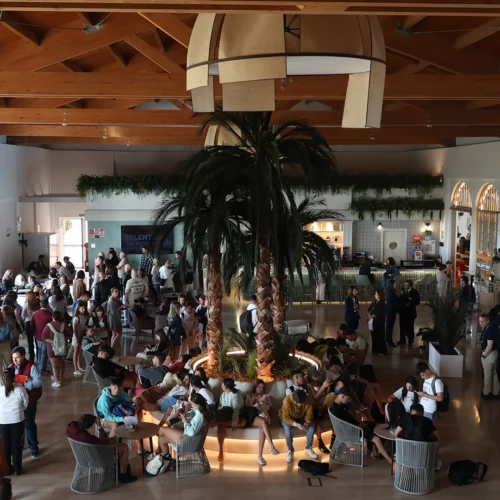 Vista general de un espacio interior amplio y luminoso con techo de madera. En el centro hay una zona circular con palmeras y asientos alrededor. Numerosas personas se encuentran sentadas, de pie o conversando en grupos distribuidos por toda la sala.