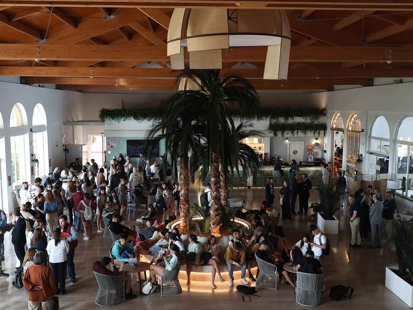 Vista general de un espacio interior amplio y luminoso con techo de madera. En el centro hay una zona circular con palmeras y asientos alrededor. Numerosas personas se encuentran sentadas, de pie o conversando en grupos distribuidos por toda la sala.