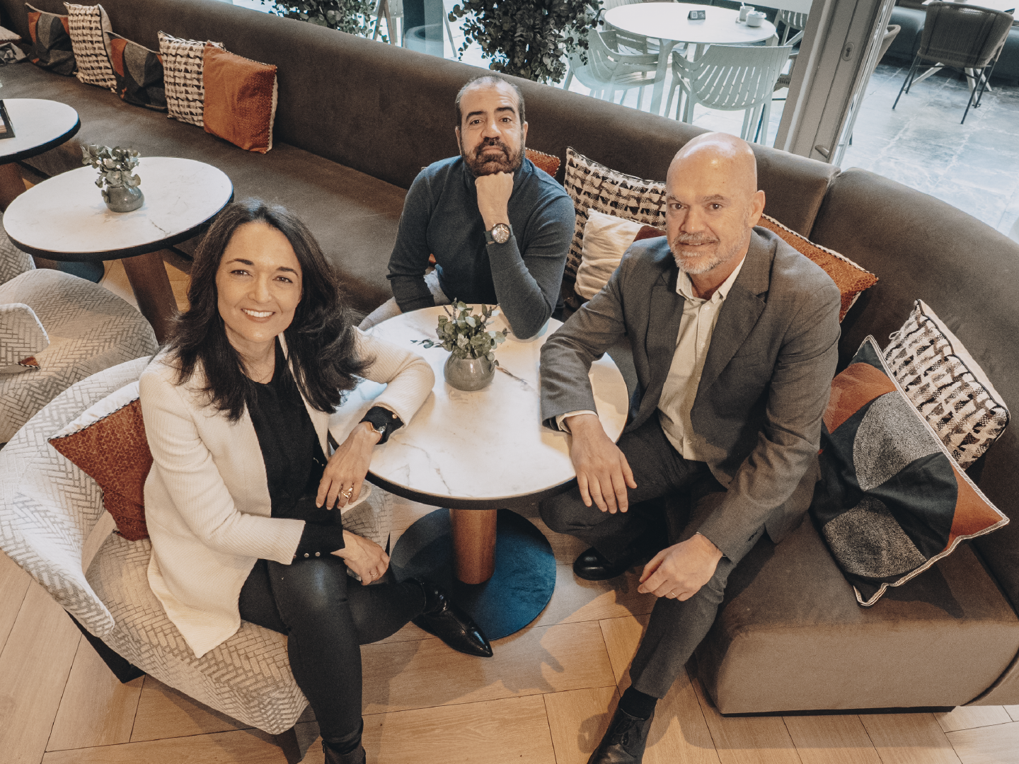 Grupo de tres personas sentadas alrededor de dos mesas redondas blancas en un espacio interior tipo lounge o cafetería moderna. El mobiliario incluye sofás tapizados en tonos beige y marrón, con cojines decorativos en colores neutros y tierra. Las personas visten ropa formal o semiformal y apoyan los brazos sobre las mesas, donde hay pequeñas plantas en maceta. El suelo es de madera clara colocada en espiga. Al fondo se aprecia un ventanal que deja entrar luz natural y una terraza exterior con mesas y sillas. El ambiente es tranquilo, cuidado y profesional.