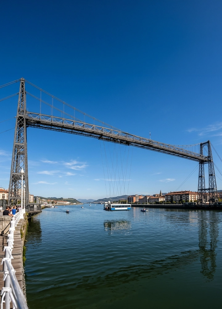 Puente metálico de gran estructura que cruza un río ancho y tranquilo, con dos torres altas unidas por una viga superior y cables verticales que sostienen una plataforma suspendida sobre el agua. En el río navegan pequeñas embarcaciones y una barcaza situada bajo el puente. A la izquierda, un paseo peatonal con barandilla blanca y varias personas caminando junto a la orilla. Al fondo aparecen edificios bajos y colinas suaves. El cielo es despejado y de un azul intenso, con luz diurna clara que se refleja en el agua.