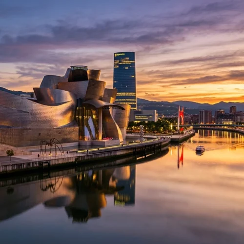 Vista panorámica de una ciudad al atardecer junto a una ría tranquila. En primer plano aparece el agua lisa, que refleja los tonos dorados y rosados del cielo y las luces de la orilla. A la izquierda destaca un gran edificio de formas curvas y revestimiento metálico, con superficies onduladas que captan la luz del ocaso. A lo largo de la ría se extiende un paseo urbano iluminado, con farolas y edificios modernos. En el centro se eleva una torre alta y rectangular con numerosas ventanas encendidas. Más al fondo se distinguen otros edificios y un puente iluminado en tonos rojizos. El paisaje urbano está rodeado por colinas oscuras, que enmarcan la escena. El cielo combina nubes suaves con colores cálidos y azules profundos, creando un contraste marcado entre la luz natural del atardecer y la iluminación artificial de la ciudad.
