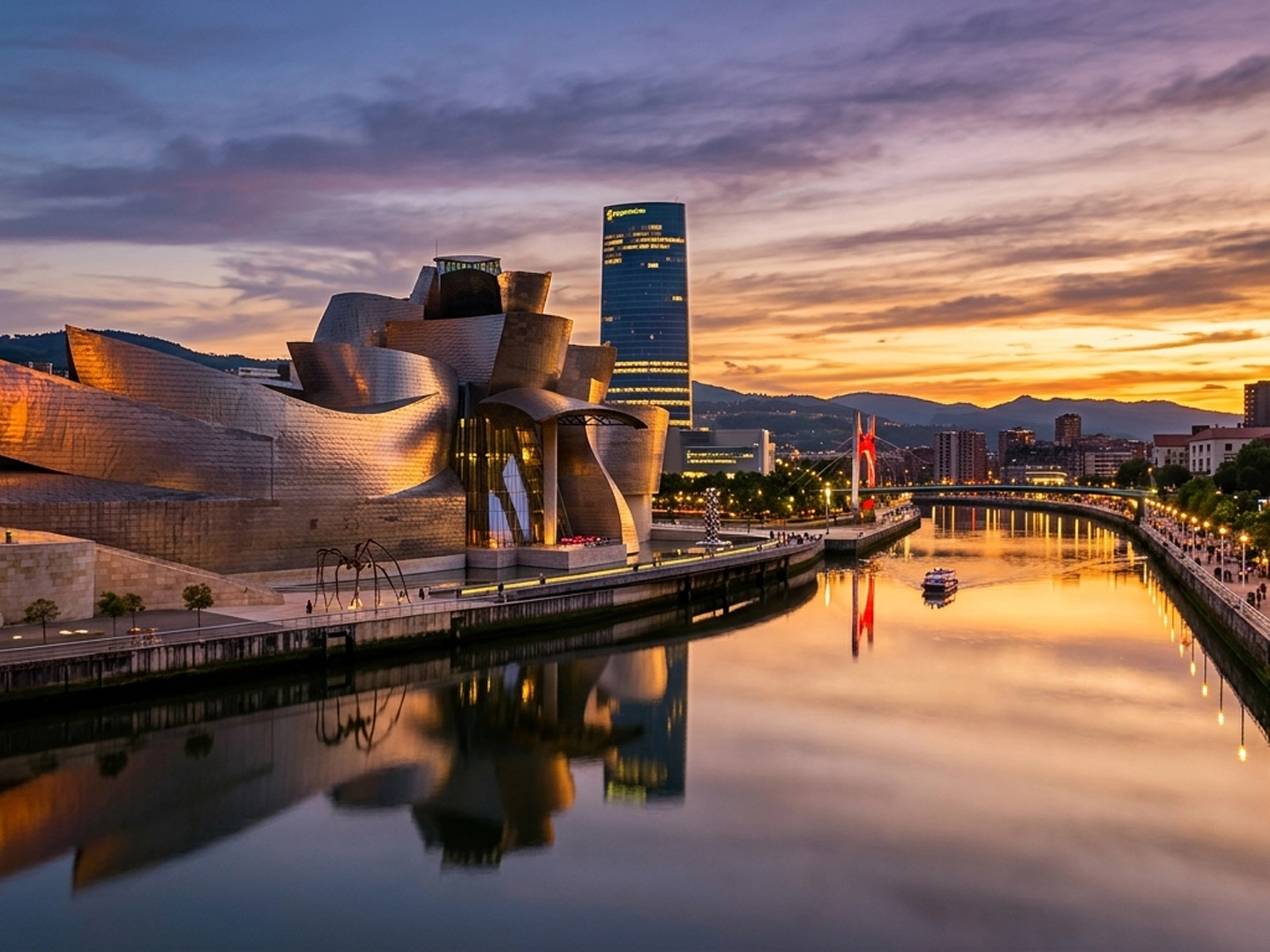 Vista panorámica de una ciudad al atardecer junto a una ría tranquila. En primer plano aparece el agua lisa, que refleja los tonos dorados y rosados del cielo y las luces de la orilla. A la izquierda destaca un gran edificio de formas curvas y revestimiento metálico, con superficies onduladas que captan la luz del ocaso. A lo largo de la ría se extiende un paseo urbano iluminado, con farolas y edificios modernos. En el centro se eleva una torre alta y rectangular con numerosas ventanas encendidas. Más al fondo se distinguen otros edificios y un puente iluminado en tonos rojizos. El paisaje urbano está rodeado por colinas oscuras, que enmarcan la escena. El cielo combina nubes suaves con colores cálidos y azules profundos, creando un contraste marcado entre la luz natural del atardecer y la iluminación artificial de la ciudad.