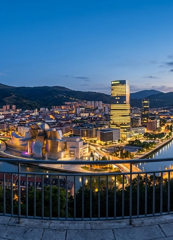 Vista panorámica urbana al atardecer desde un mirador con barandilla metálica en primer plano. En la parte inferior izquierda destaca un edificio de formas curvas y revestimiento metálico junto a una ría que atraviesa la ciudad. A lo largo del agua se alinean calles iluminadas y edificios modernos y tradicionales. En el centro sobresale una torre alta de planta rectangular con muchas ventanas encendidas. Al fondo se elevan colinas oscuras que rodean la ciudad. El cielo es azul profundo, con luz de anochecer que contrasta con la iluminación artificial urbana.