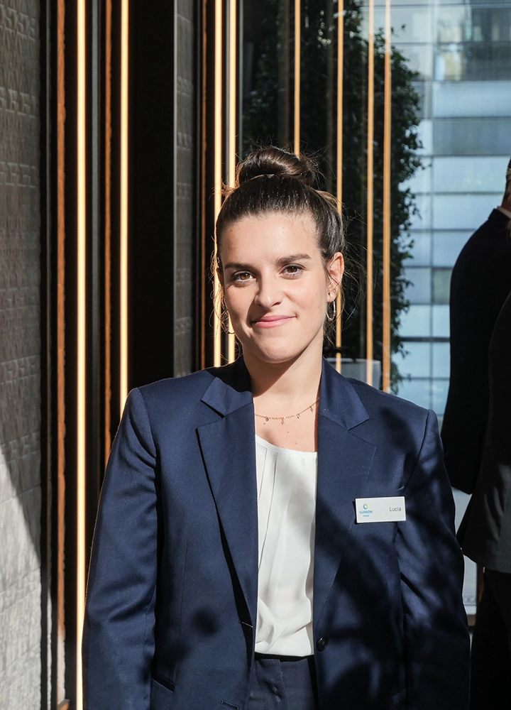Persona situada en la entrada o zona de paso de un edificio contemporáneo, con iluminación natural lateral. Viste un traje de chaqueta azul oscuro con una blusa clara y una placa identificativa en la solapa. El rostro está desenfocado. Detrás se observan paneles verticales oscuros alternados con líneas de luz cálida, creando un efecto elegante. A través de un ventanal se distinguen edificios urbanos y vegetación.