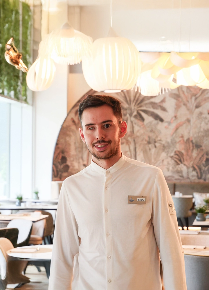 Persona de pie en el interior de un restaurante moderno y luminoso. Lleva una camisa blanca de manga larga, cerrada hasta el cuello, con una placa identificativa rectangular en el pecho. El rostro aparece desenfocado para preservar la privacidad. El espacio tiene mesas y sillas de diseño contemporáneo, tonos claros y materiales naturales. Del techo cuelgan varias lámparas grandes de formas orgánicas y color blanco que aportan una luz cálida. Al fondo se aprecia una pared decorativa con un mural de motivos vegetales y un arco que estructura el espacio.