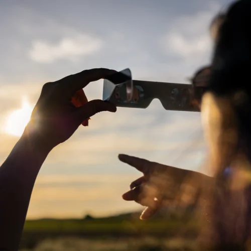 Persona en primer plano observada desde detrás, alzando una mano que sostiene unas gafas especiales para eclipses. Delante, otra mano señala hacia el cielo. El sol está bajo, cerca del horizonte, parcialmente cubierto por las gafas, y emite una luz dorada que atraviesa el paisaje. Al fondo se extiende un campo abierto con vegetación baja y un cielo amplio con nubes suaves, iluminado por la luz cálida del atardecer.