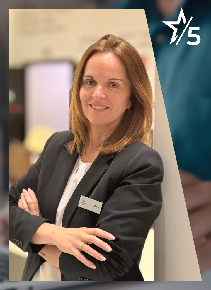 Persona adulta en plano medio, con el rostro difuminado, apoyada ligeramente sobre un panel vertical claro. Viste chaqueta oscura sobre blusa clara y lleva una acreditación con nombre visible en el pecho. Los brazos están cruzados a la altura del torso. El fondo es interior y desenfocado, con tonos cálidos. En la esquina superior derecha aparece un elemento gráfico con una estrella y el número “5” en blanco sobre fondo azul oscuro.