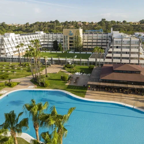 Vista aérea de un complejo hotelero rodeado de jardines. En primer plano hay una gran piscina exterior de agua azul, con palmeras y zonas de césped alrededor. A un lado se sitúa un edificio bajo con terraza, y al fondo se alza el edificio principal del hotel, de varias plantas y fachadas claras, con caminos y áreas verdes bien distribuidas bajo luz natural de día.
