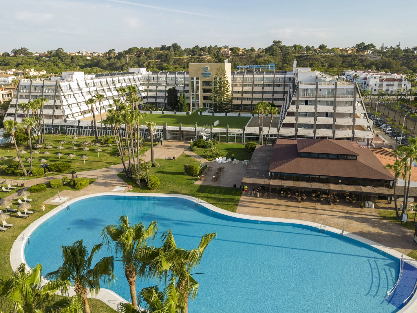 Vista aérea de un complejo hotelero rodeado de jardines. En primer plano hay una gran piscina exterior de agua azul, con palmeras y zonas de césped alrededor. A un lado se sitúa un edificio bajo con terraza, y al fondo se alza el edificio principal del hotel, de varias plantas y fachadas claras, con caminos y áreas verdes bien distribuidas bajo luz natural de día.