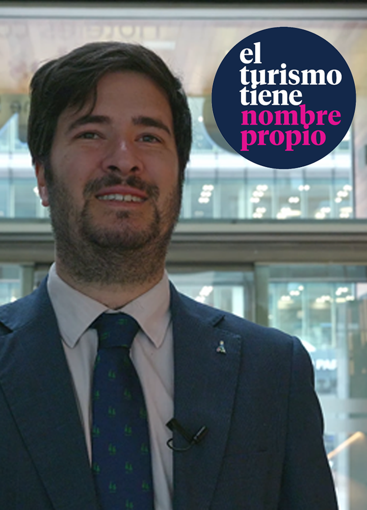 Plano medio de una persona adulta vestida con traje azul oscuro, camisa clara y corbata verde con pequeños motivos. El rostro aparece completamente desenfocado. En la solapa de la chaqueta hay un pequeño pin y un micrófono de clip, lo que sugiere una intervención o entrevista. El fondo es un espacio interior moderno con grandes superficies acristaladas y luz artificial, que deja ver estructuras y puntos de luz desenfocados. En la parte superior derecha aparece un círculo azul con el texto “el turismo tiene nombre propio”, donde las palabras “el turismo tiene” están en blanco y “nombre propio” en color rosa.