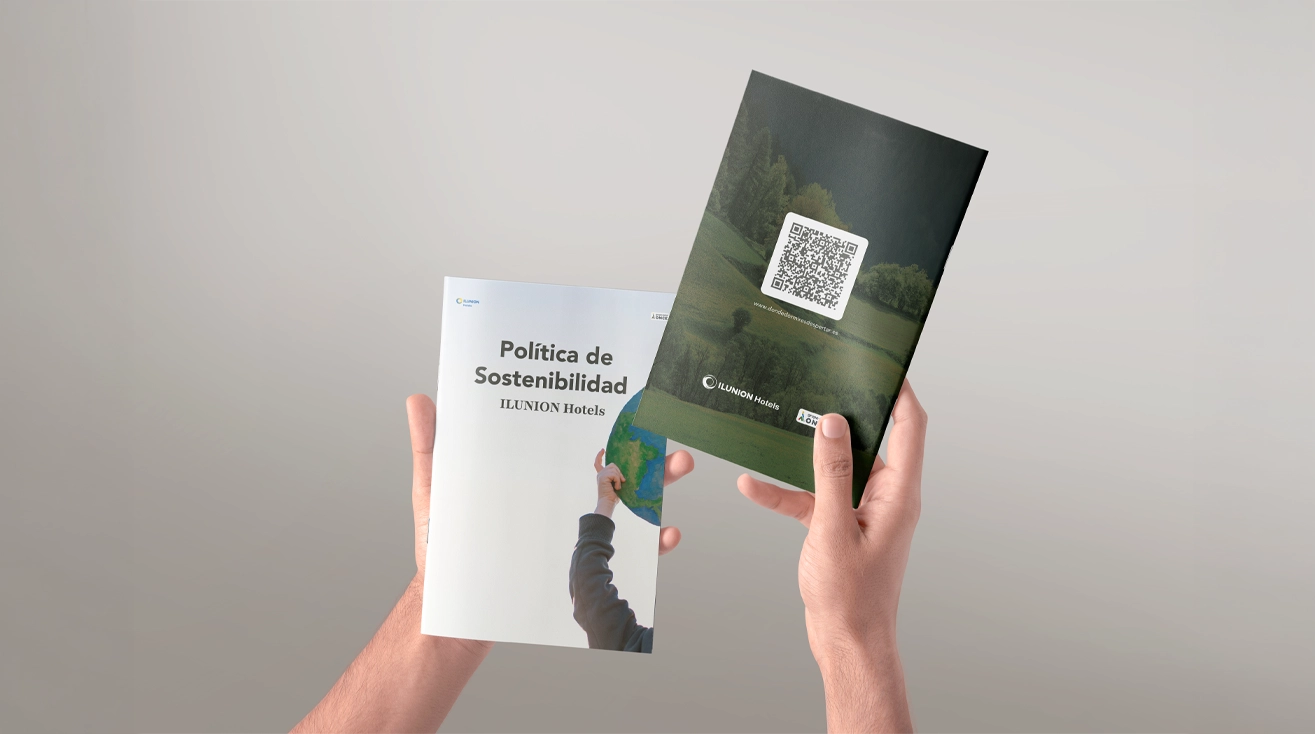 Dos manos sostienen dos folletos a la altura del pecho, recortadas sobre un fondo liso en tono gris claro. En la mano izquierda hay un folleto blanco con el título “Política de Sostenibilidad” y el subtítulo “ILUNION Hotels”. En la portada aparece una imagen parcial de una persona levantando un globo terráqueo con una mano, visible desde el borde inferior. En la mano derecha hay un folleto de portada oscura, en tonos verdes y negros, con una imagen de paisaje natural desenfocada. En la parte superior de la portada destaca un código QR blanco y, en la zona inferior, el logotipo y el texto “ILUNION Hotels”. La composición es limpia y centrada, con iluminación suave y uniforme, transmitiendo una imagen corporativa y relacionada con sostenibilidad y comunicación institucional.