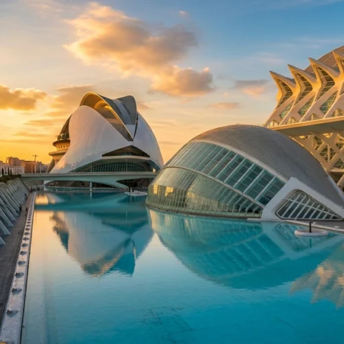 Complejo arquitectónico contemporáneo con edificios blancos de formas curvas y futuristas, rodeados por un gran estanque de agua azul turquesa que refleja las estructuras. En primer plano, el agua tranquila ocupa casi toda la superficie, delimitada por una pasarela con elementos repetidos en forma de arcos inclinados. A la derecha se alza una construcción alargada con estructura reticulada y huecos triangulares. Al fondo, un edificio con cubierta curva y volúmenes superpuestos destaca contra un cielo con nubes suaves iluminadas por la luz cálida del atardecer. El conjunto transmite amplitud, simetría y modernidad.