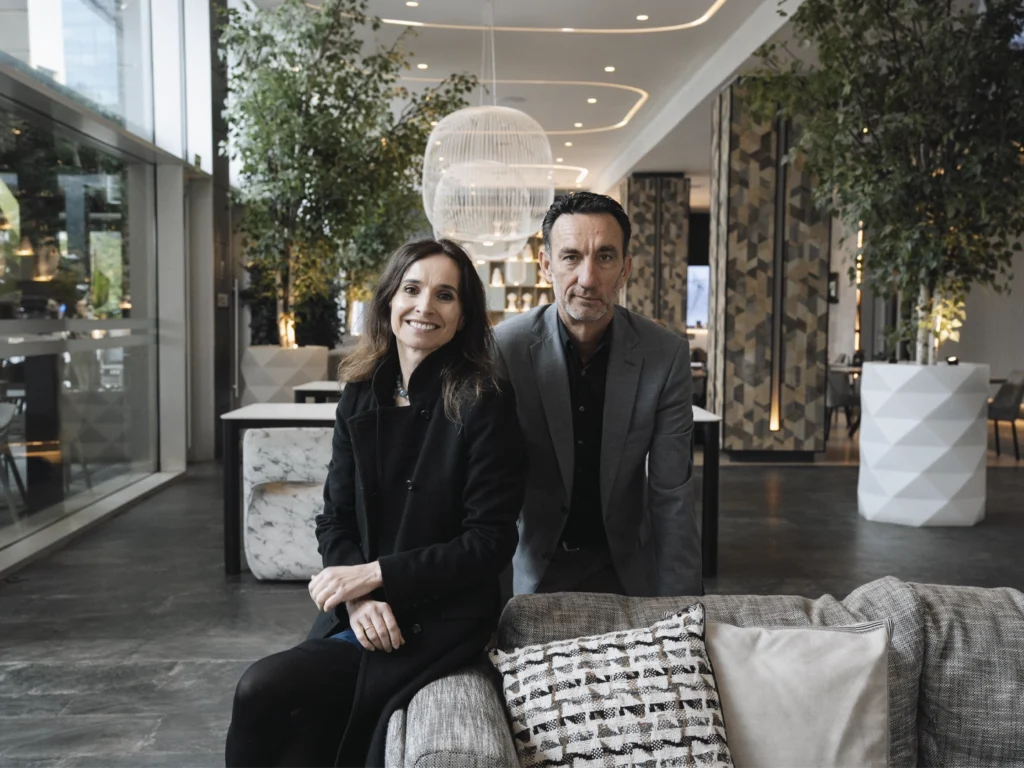 Espacio interior amplio y luminoso, similar al vestíbulo de un hotel moderno, con grandes ventanales, plantas altas en macetas y elementos decorativos contemporáneos. En primer plano, dos personas adultas están sentadas juntas en un sofá tapizado, vestidas con ropa formal de tonos oscuros, apoyadas ligeramente hacia delante. Delante del sofá hay cojines decorativos y, al fondo, se aprecian lámparas colgantes, columnas con revestimientos geométricos y una zona de paso abierta que aporta profundidad al entorno.
