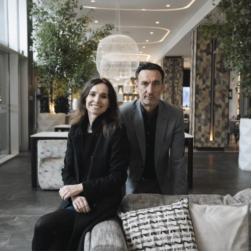 Espacio interior amplio y luminoso, similar al vestíbulo de un hotel moderno, con grandes ventanales, plantas altas en macetas y elementos decorativos contemporáneos. En primer plano, dos personas adultas están sentadas juntas en un sofá tapizado, vestidas con ropa formal de tonos oscuros, apoyadas ligeramente hacia delante. Delante del sofá hay cojines decorativos y, al fondo, se aprecian lámparas colgantes, columnas con revestimientos geométricos y una zona de paso abierta que aporta profundidad al entorno.