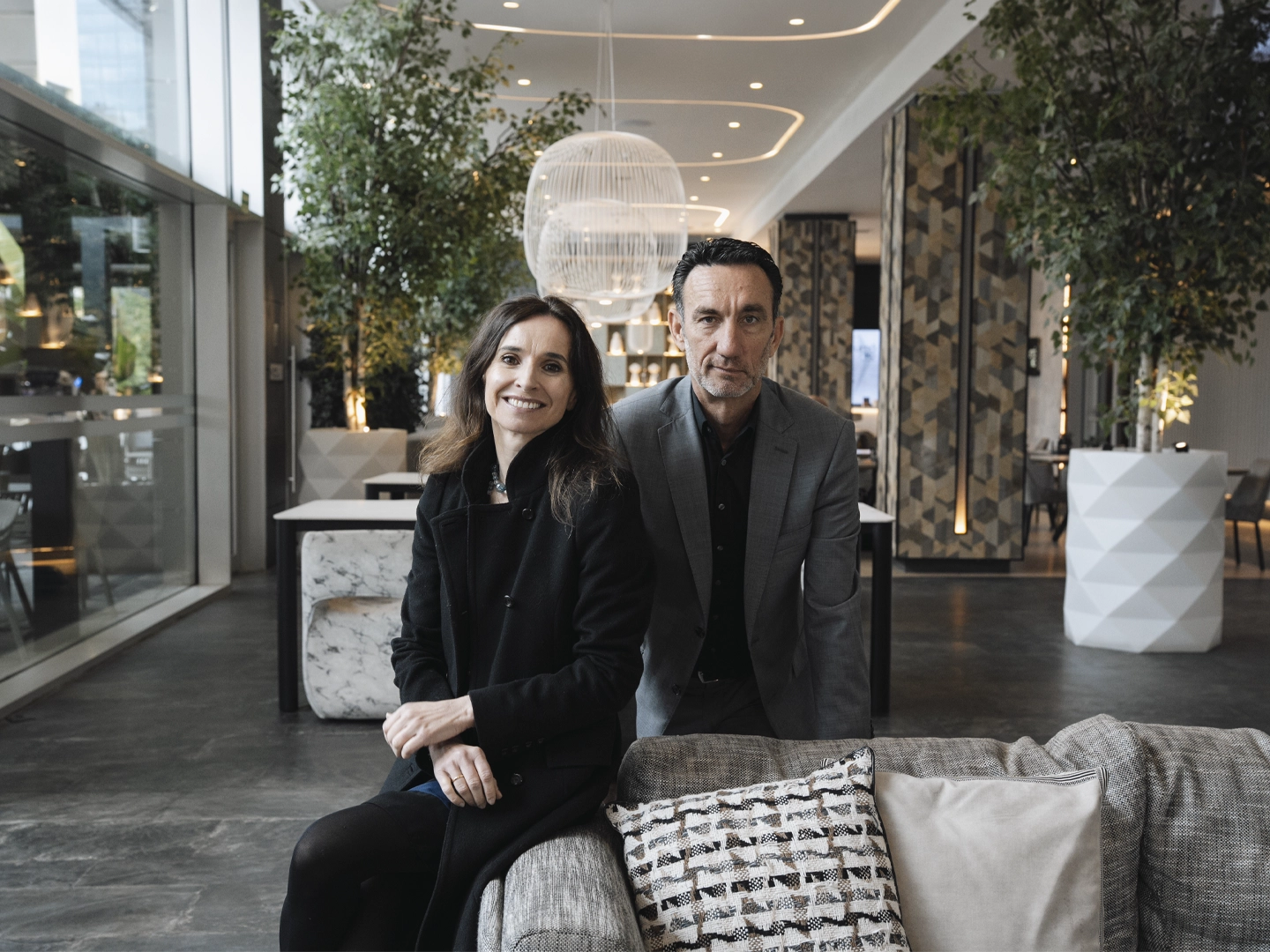 Espacio interior amplio y luminoso, similar al vestíbulo de un hotel moderno, con grandes ventanales, plantas altas en macetas y elementos decorativos contemporáneos. En primer plano, dos personas adultas están sentadas juntas en un sofá tapizado, vestidas con ropa formal de tonos oscuros, apoyadas ligeramente hacia delante. Delante del sofá hay cojines decorativos y, al fondo, se aprecian lámparas colgantes, columnas con revestimientos geométricos y una zona de paso abierta que aporta profundidad al entorno.