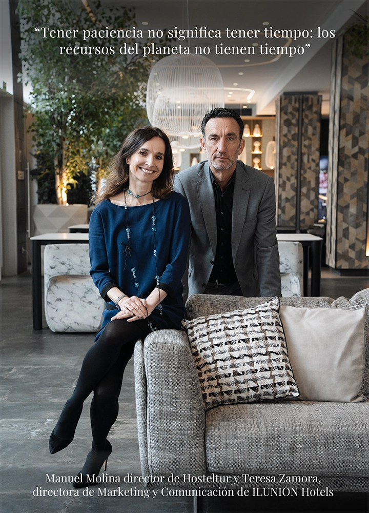 Dos personas adultas sentadas juntas en un sofá tapizado dentro de un espacio interior amplio y contemporáneo, similar al vestíbulo de un hotel, con grandes ventanales, luz natural, plantas de interior, lámparas colgantes y elementos decorativos modernos al fondo. Ambas llevan vestimenta formal en tonos neutros y oscuros, y en primer plano se aprecian cojines decorativos sobre el sofá. En la parte superior de la imagen aparece una cita en texto blanco que dice: “Tener paciencia no significa tener tiempo: los recursos del planeta no tienen tiempo”. En la parte inferior se incluye el texto: “Manuel Molina Director de Hosteltur y Teresa Zamora, directora de Marketing y Comunicación de ILUNION Hotels”.